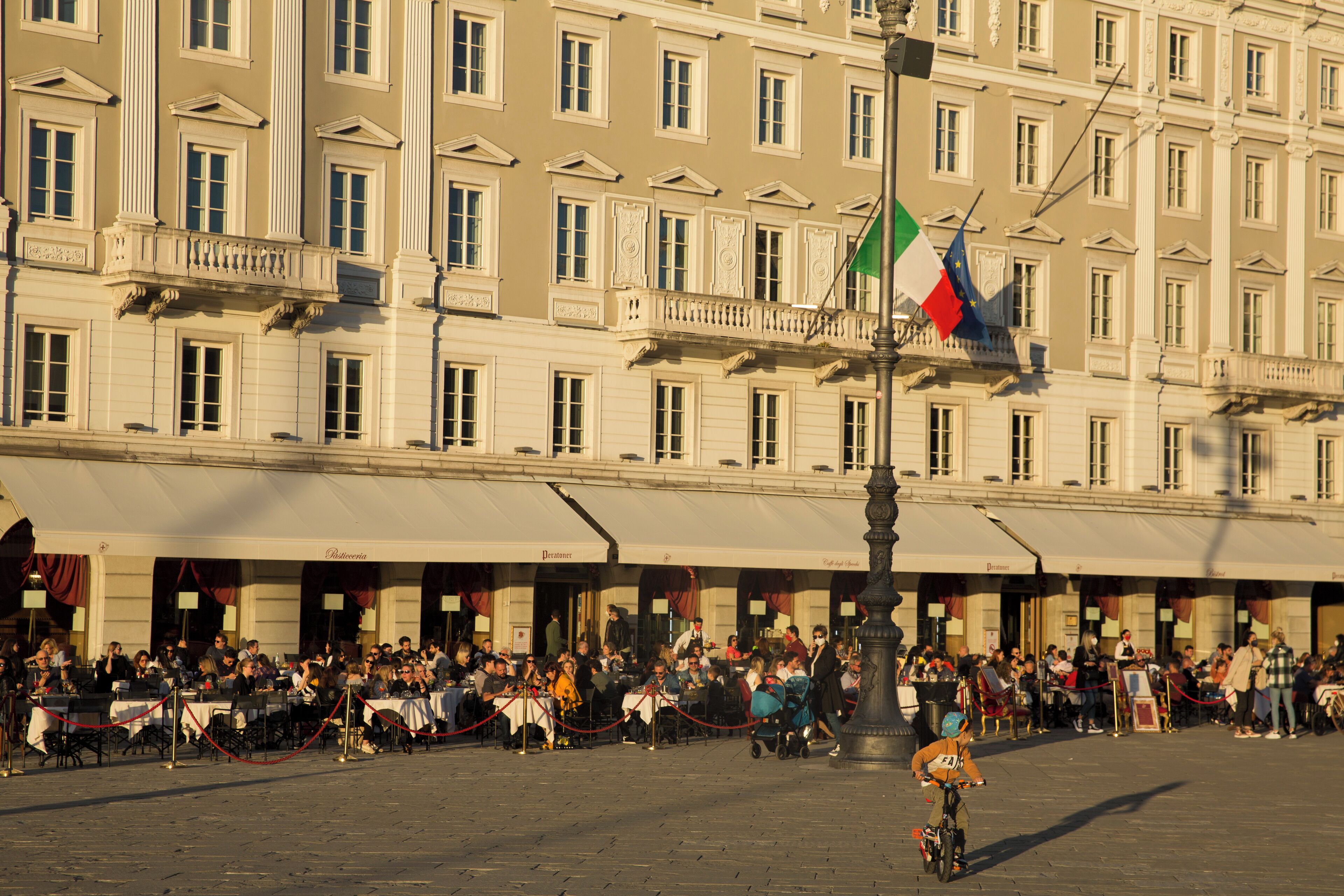 A public square with a crowd of people, a cyclist, and a flagpole with flags.