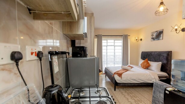 Ein kompaktes Hotelzimmer mit einer Küchenzeile, einem Bett mit Kopfteil, einem Wasserspender und einem großen Fenster mit Vorhängen.