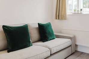 A beige sofa with two dark green cushions in a room with a white radiator and a window with curtains.