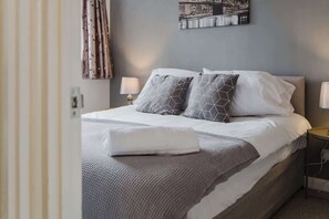 Et hotelværelse med en seng, to sengeborde med lamper, et bybillede på væggen og et gardin.