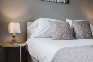 A neatly made bed with white bedding and two patterned pillows, a bedside lamp, and a small round table with a lamp.