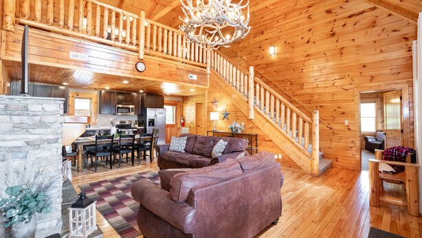 A spacious log cabin interior with a large wooden staircase, a cozy living area with a brown leather sofa, a dining area, and a kitchen. - Luxury Cabin w/Indoor Pool, Mini Golf & Mtn Views (Sevierville)