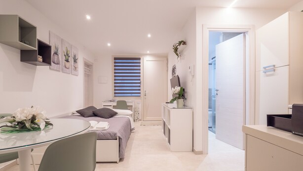 Un dormitorio moderno y bien iluminado con cama, mesita de noche y escritorio con silla. Hay una pequeña zona de estar con mesa y sillas, y un baño visible a través de una puerta abierta.