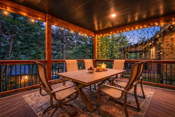 Un área de comedor al aire libre techada, con una mesa y sillas, rodeada por una terraza y barandilla de madera, con luces colgantes y vegetación al fondo.