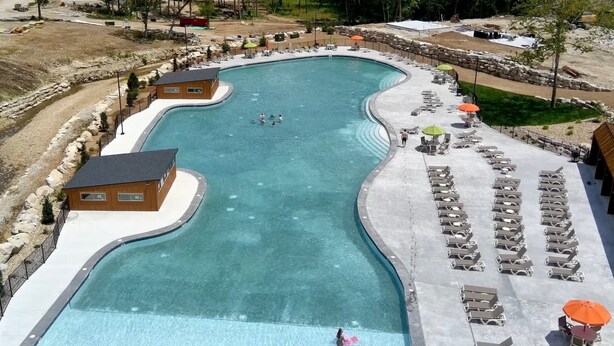 Une grande piscine incurvée, dotée de chaises longues et de parasols, entourée d’une terrasse en béton et d’un petit bâtiment.