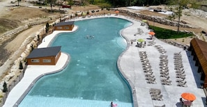 Una gran piscina curada con sillas de descanso y sombrillas, rodeada por una terraza de hormigón y un pequeño edificio.