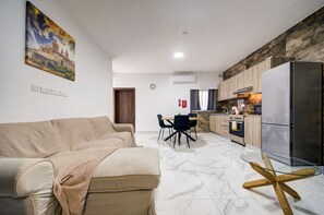 Ein modernes Wohnzimmer mit einer Couch, einem Essbereich mit Stühlen, einer Küche inklusive Kühlschrank und Herd sowie einem Bild an der Wand.