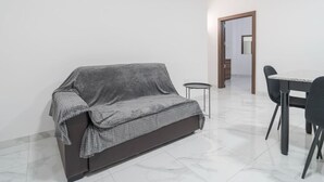 A modern living room with a grey sofa, a small round table, and a dining area with a white table and black chairs. - Charming 1BR with comfy bed w/WIFI & AC in Tarxien (Tarxien)