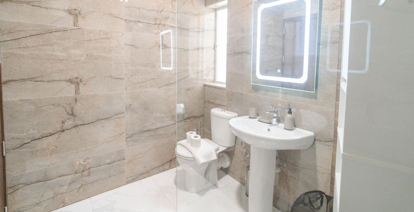 Une salle de bain moderne avec un mur en marbre, une cabine de douche en verre, des toilettes et un lavabo surmonté d’un miroir.