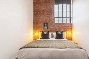 Ein Bett mit Kopfteil, eine Ziegelwand, zwei Wandleuchten und ein Fenster mit Gittermuster.