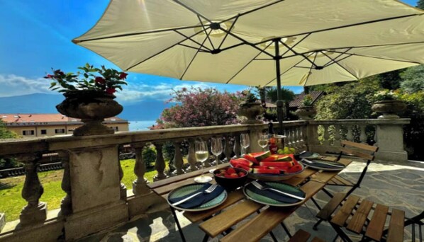 An outdoor dining area with a table set for two, a large umbrella, and a view of a garden and distant buildings.