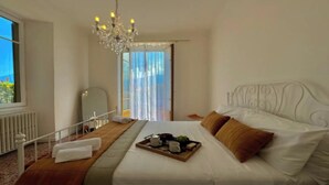 A hotel room with a large bed, a chandelier, a window with curtains, and a tray with breakfast items. - A Hidden Gem in Menaggio – Elegant Apartment with Terrace & Lake Views (Menaggio)