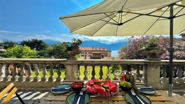 A terrace with a table set for a meal, a large umbrella, and a view of a building and trees. - A Hidden Gem in Menaggio – Elegant Apartment with Terrace & Lake Views (Menaggio)