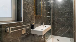 Een moderne badkamer met een marmeren wand, een witte wastafel, een glazen douchecabine en een spiegel met gouden lijst.