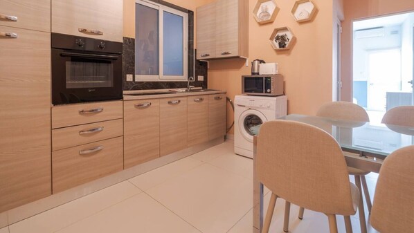 A compact kitchen with a built-in oven, microwave, and washing machine, a dining area with a table and chairs, and a window providing natural light.