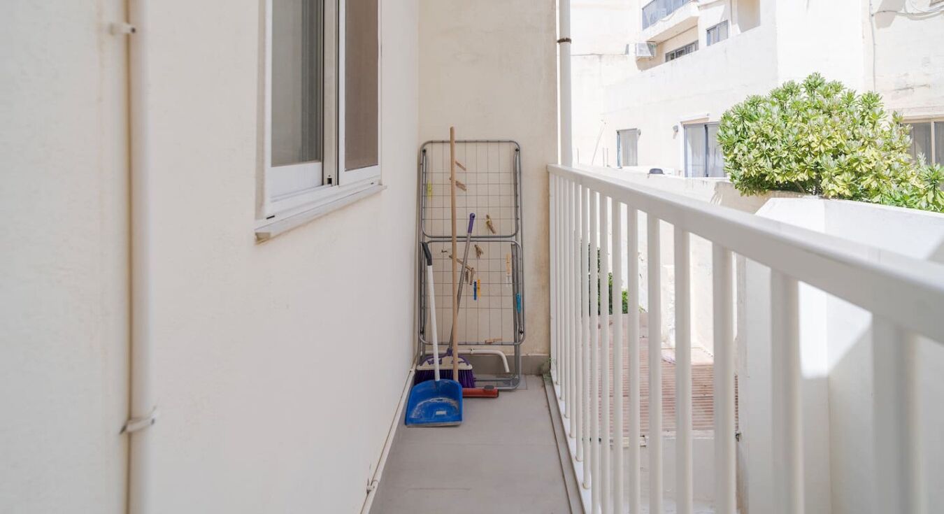 Un balcón estrecho con barandilla blanca, una ventana y un tendedero metálico con utensilios de limpieza.