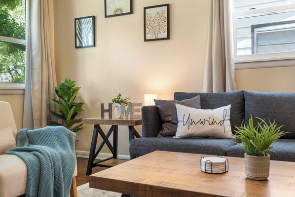 A cozy living room with a grey sofa, a wooden coffee table, and a side table with a plant and a sign that reads "HOME". - Chic Bungalow Private Gym + Office Weekly & Monthly Near Bass Pro & Branson" (Springfield)