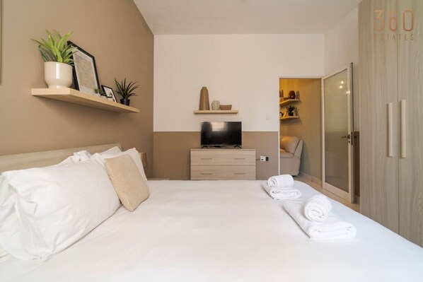 A hotel room with a bed, a TV on a dresser, a closet, and a shelf with plants.