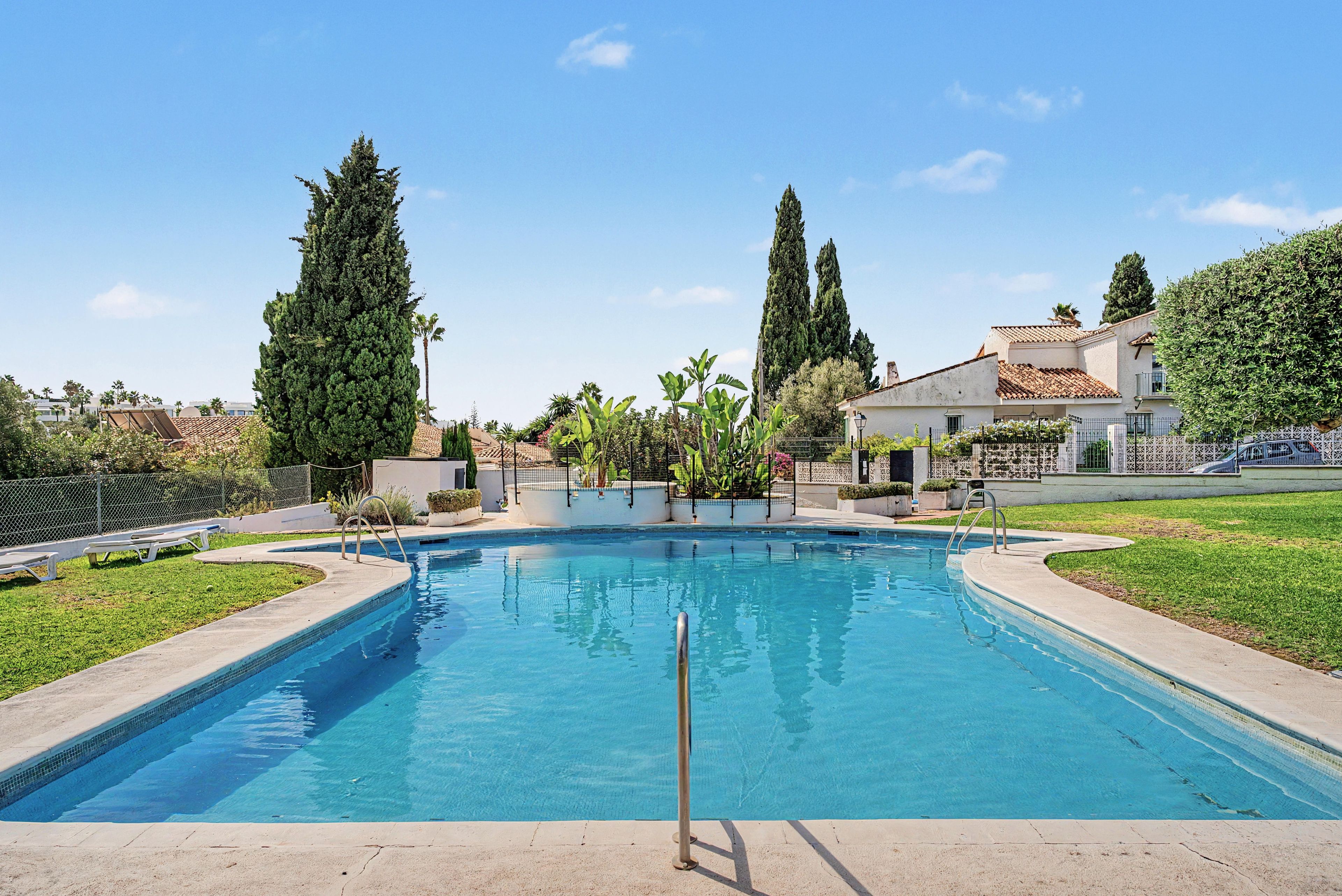 Ein Swimmingpool mit glatter, blauer Wasseroberfläche, umgeben von einem weißen Zaun sowie Bäumen und Gebäuden im Hintergrund.