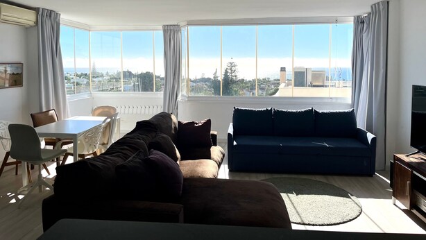 A living room with a sofa, a dining table with chairs, and a large window offering a city view. - Bright 'Luminoso Marbella' Apartment with Sea Views, Wi-Fi and Air Conditioning (Marbella)