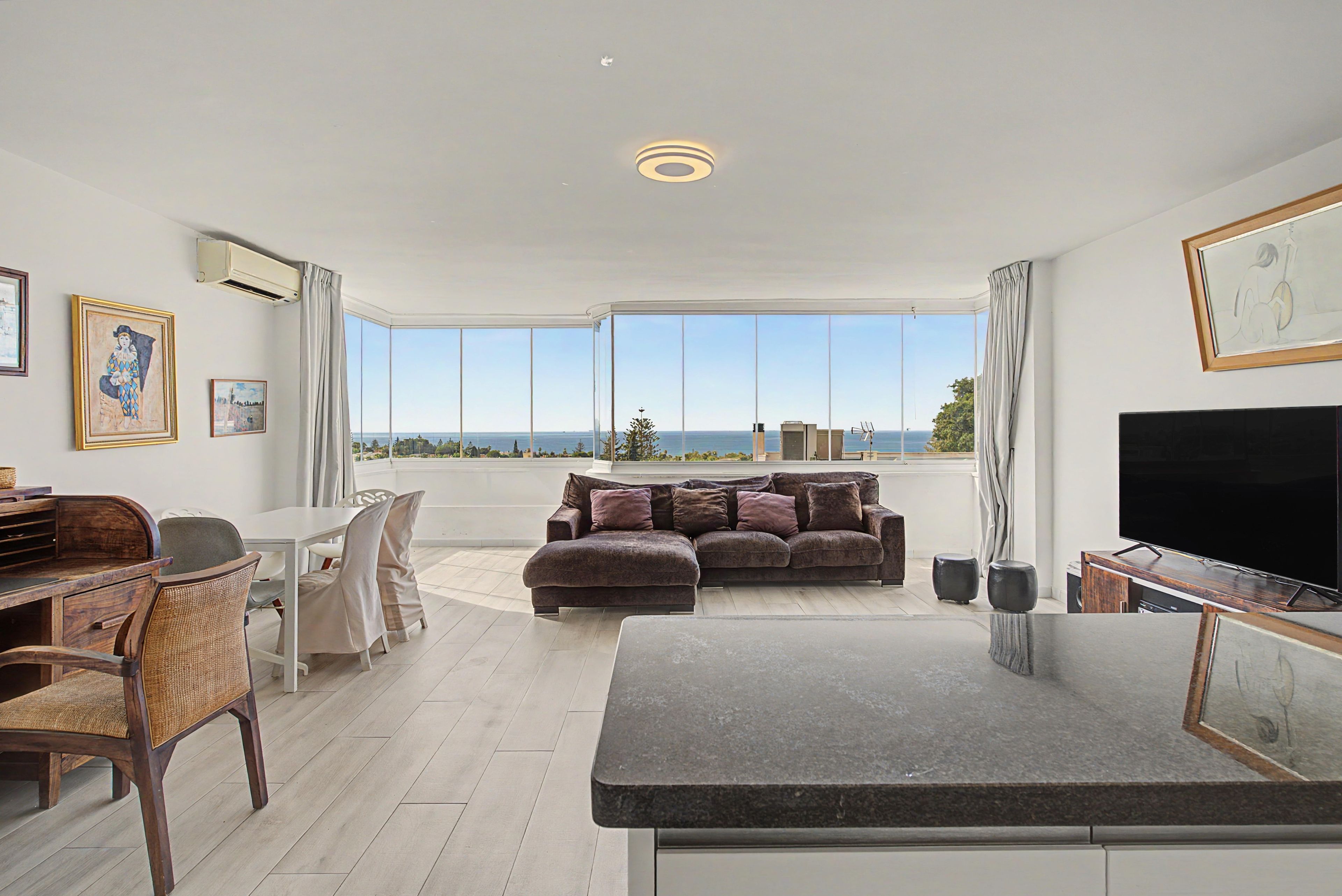 A modern living room with a large flat-screen TV, a sofa, a dining area with a table and chairs, and a view of the sea through large windows.
