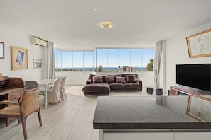 A modern living room with a large flat-screen TV, a sofa, a dining area with a table and chairs, and a view of the sea through large windows.