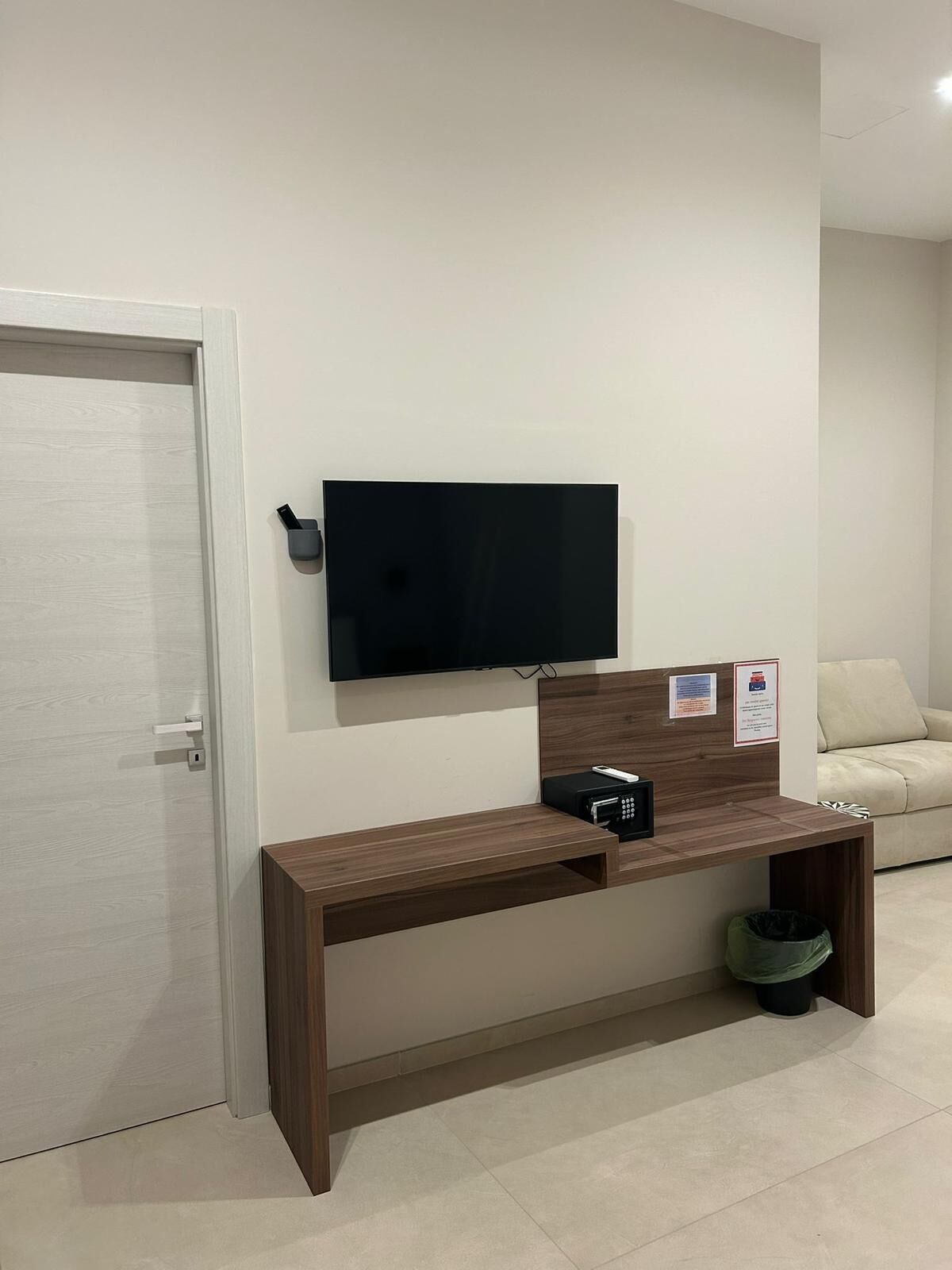 Quarto moderno com televisão de tela plana fixada na parede, escrivaninha de madeira com micro-ondas, lixeira e sofá bege.