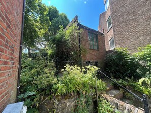 A narrow alley with brick buildings, dense foliage, and a stone wall.