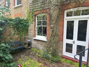 A brick house with a white door and window, a metal shelf with potted plants, a bench, and a small garden area with greenery.