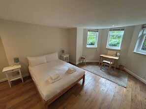 Et soveværelse med en træseng, sengeborde, et skrivebord og to vinduer med udsigt til grøn vegetation.