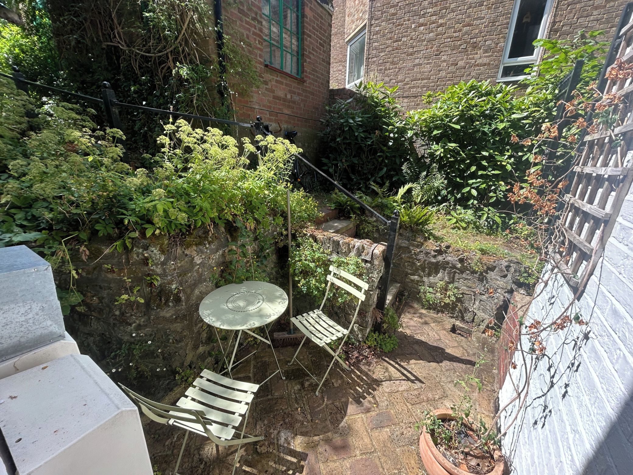 A small outdoor patio with a table and chairs, surrounded by dense foliage and a brick wall.