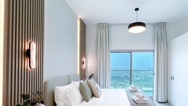 A modern bedroom with a large bed, a bedside table, a vase with a plant, and a view of the city through large windows. - Urban Escape Studio Pool, Gym & Stylish Living (Dubai)