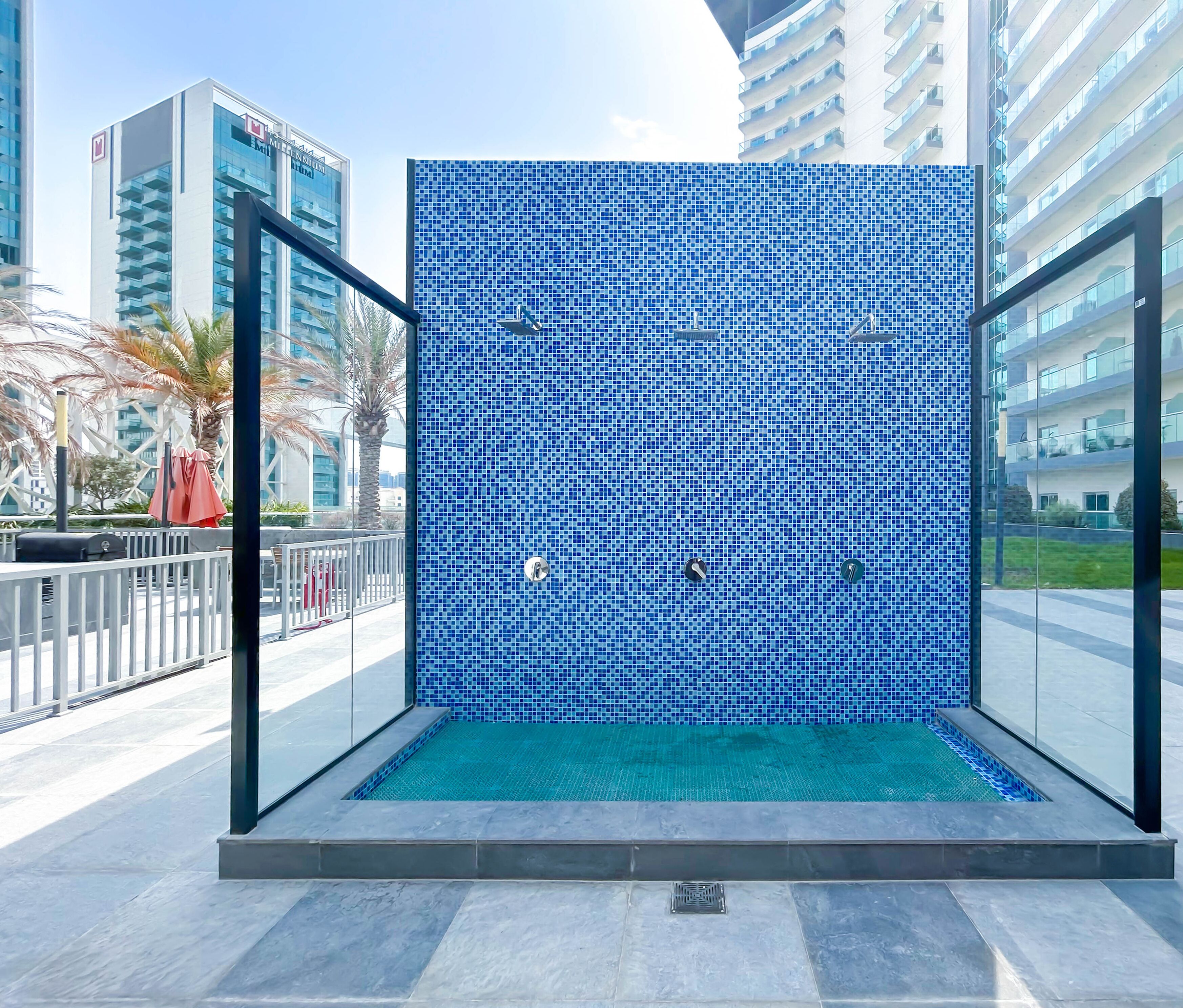 A modern outdoor pool with a blue mosaic wall, surrounded by glass railings and palm trees.
