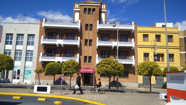 A multi-story building with a central tower, balconies, and a pedestrian area with trees and a cyclist. - Hotel Luz del Guadiana (Ayamonte)