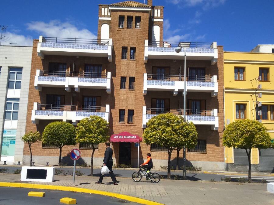 Un edificio de varios pisos con una torre central, balcones y un área peatonal con árboles y un ciclista.