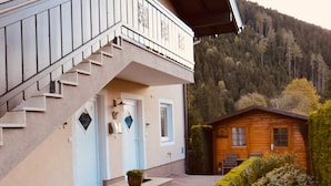 Un bâtiment à deux étages doté d’un balcon, une cabane en bois en arrière-plan et un paysage montagneux.