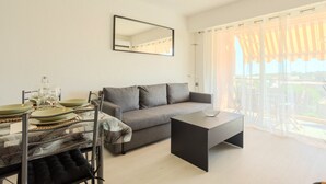En moderne stue med en grå sofa, et spisebord dækket med tallerkener og glas, et sort sofabord og et stort vindue med lette gardiner.