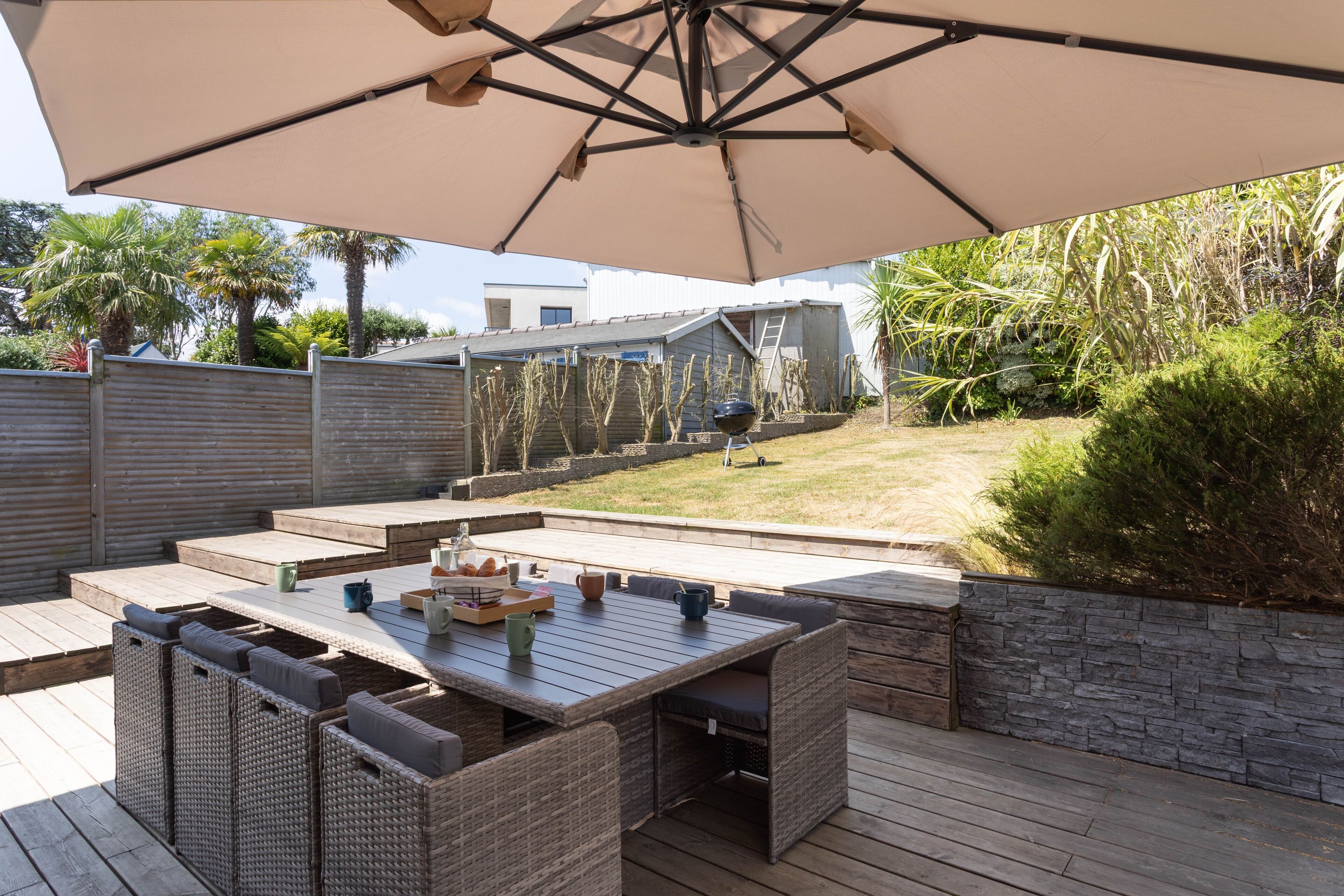 Patio esterno con tavolo e sedie, un ombrellone grande e vista su un giardino sul retro con un pontile in legno e un muro in pietra.