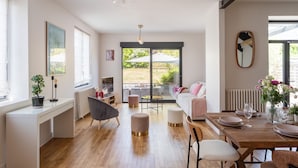 A modern living room with a wooden dining table set for four, a grey sofa, and a large window with a view of greenery.