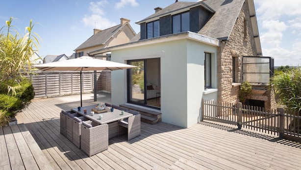 A modern house with a wooden deck, outdoor seating, and a stone chimney.