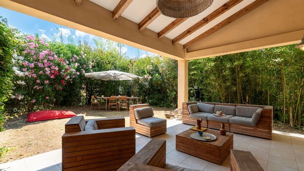 An outdoor patio with wooden furniture, a dining area, and a view of a garden with pink flowers.