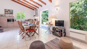 A spacious living area with a dining table, chairs, a TV, and a large window with a view of greenery.