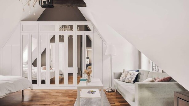 Un dormitorio tipo loft moderno con techo inclinado, piso de madera, un armario blanco, un sofá gris y una pequeña mesa blanca.