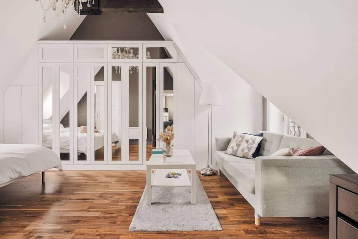 Un dormitori tipus loft modern amb sostre inclinat, terres de fusta, un armari blanc, un sofà gris i una taula petita blanca.