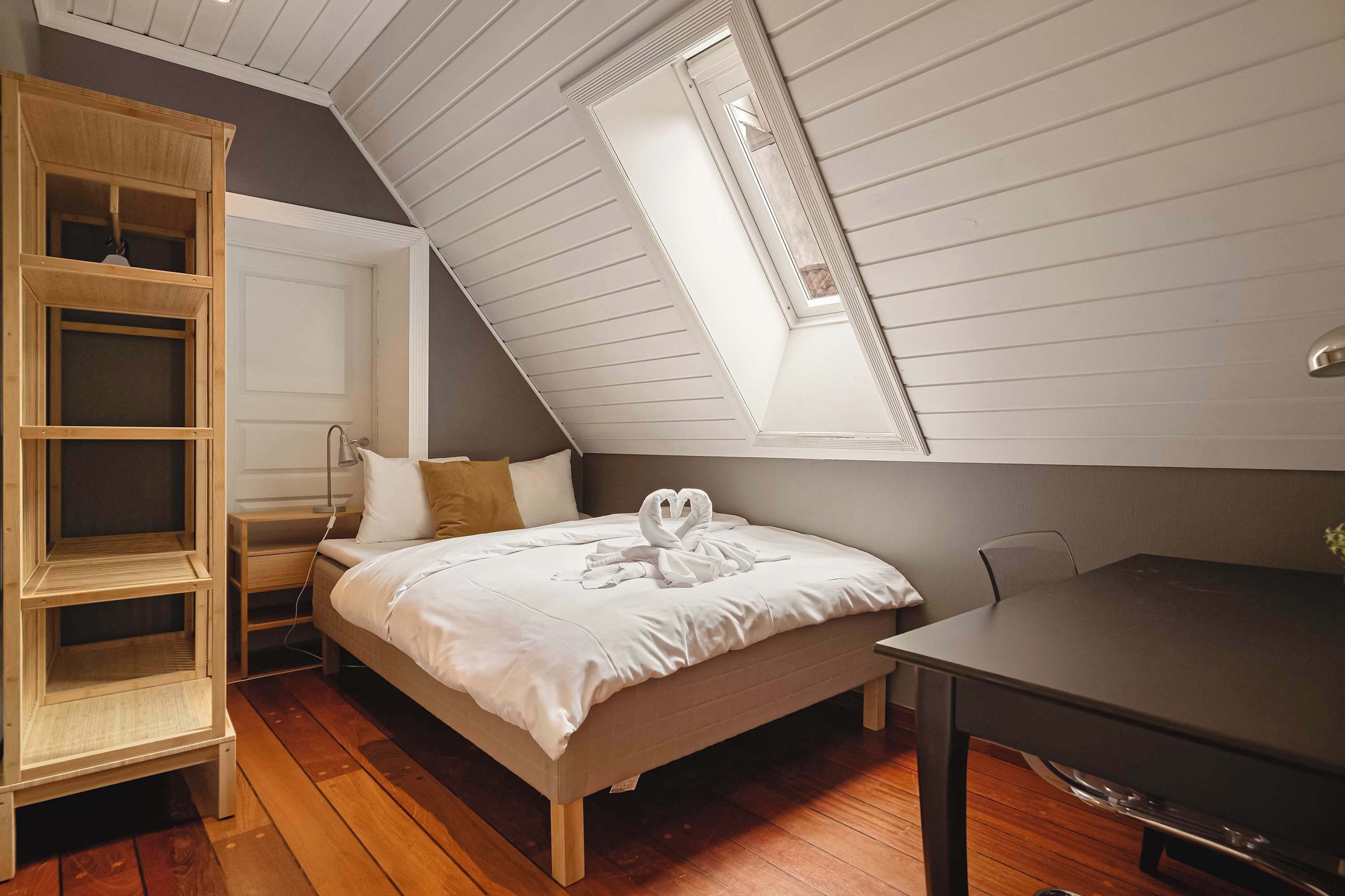 A bedroom with a sloped ceiling, a bed with white bedding, a wooden nightstand, a desk, and a built-in shelving unit.