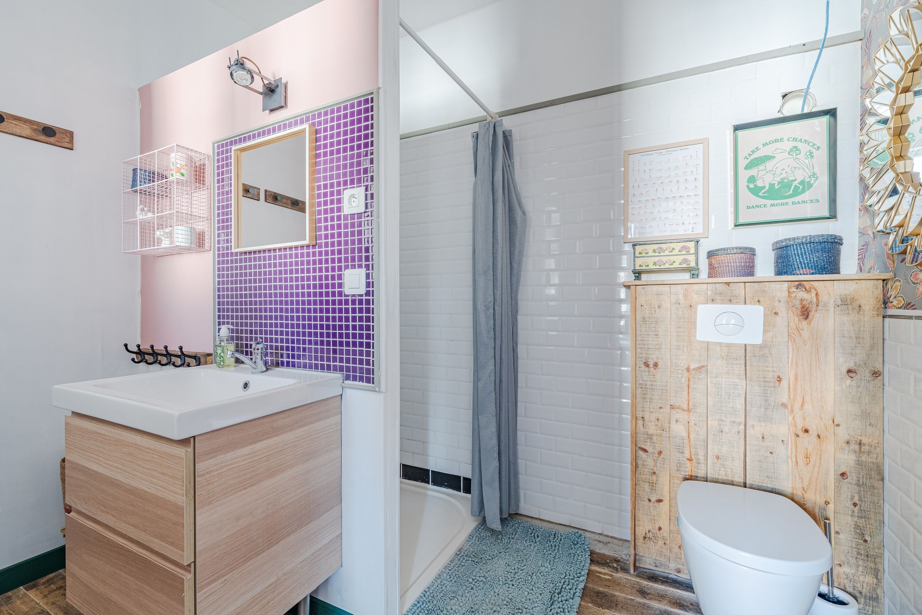 Ein Badezimmer mit weißem Waschbecken, Holzschrank, violetten Fliesen, einer Dusche und einer Toilette an einer Wandpaneele verkleideten Wand.