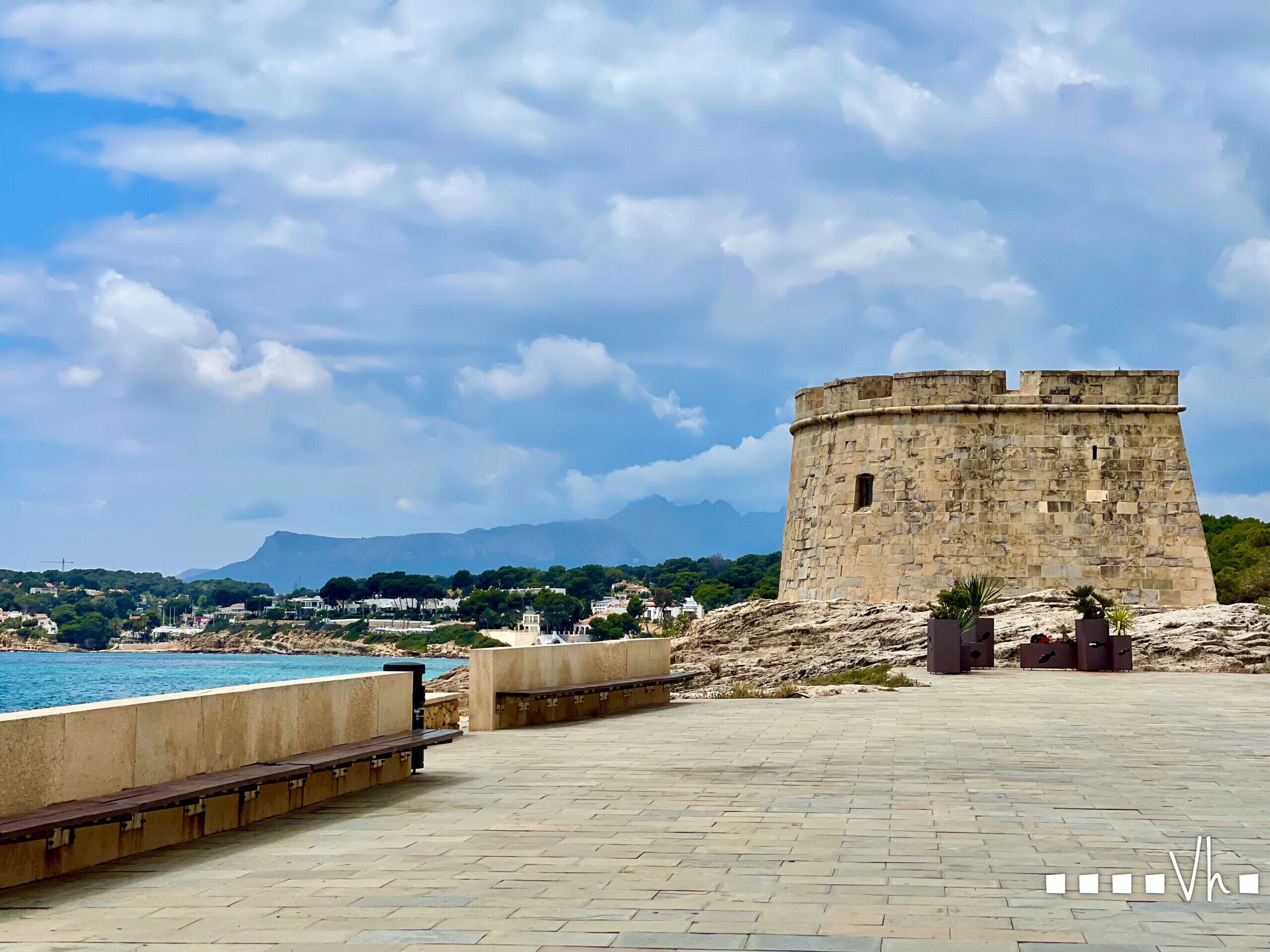 Une forteresse en pierre dominant une zone côtière, avec une promenade pavée et des bancs.