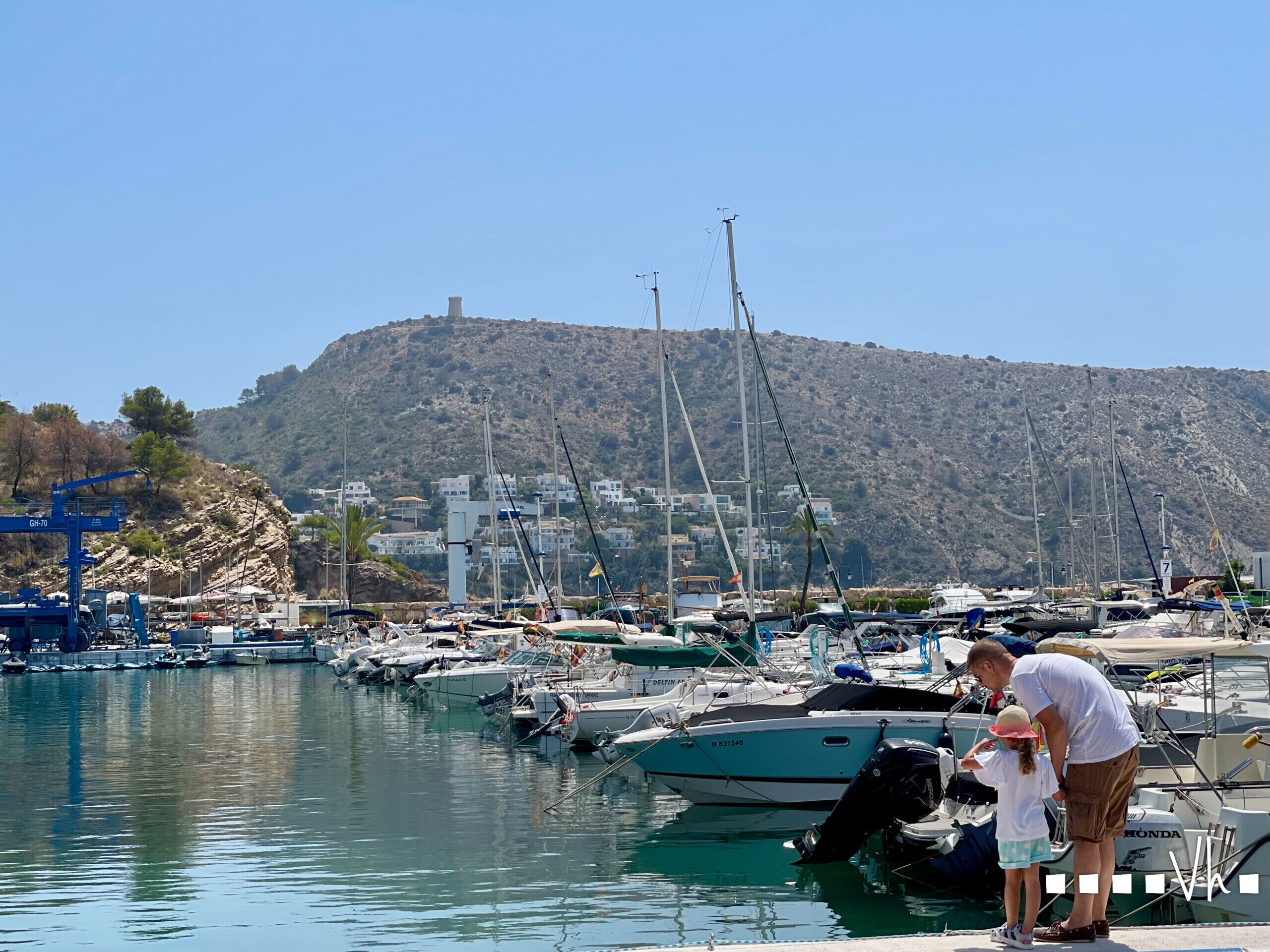 A marina with boats docked, a man and a child by the water, and a hill with buildings in the background.