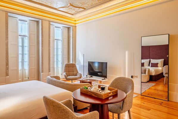 Una habitación de hotel con una cama grande, televisión, zona de estar con sillas y un espejo que refleja la habitación.