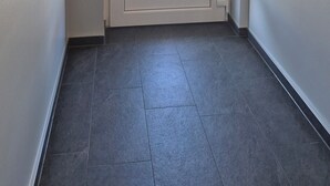 A hallway with a white door featuring a window and a handle. The floor is tiled, and the walls are plain.